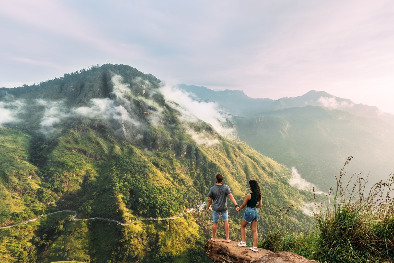 A Sri Lankan guide walking with a traveller along a footpath through tea-covered hills at sunrise